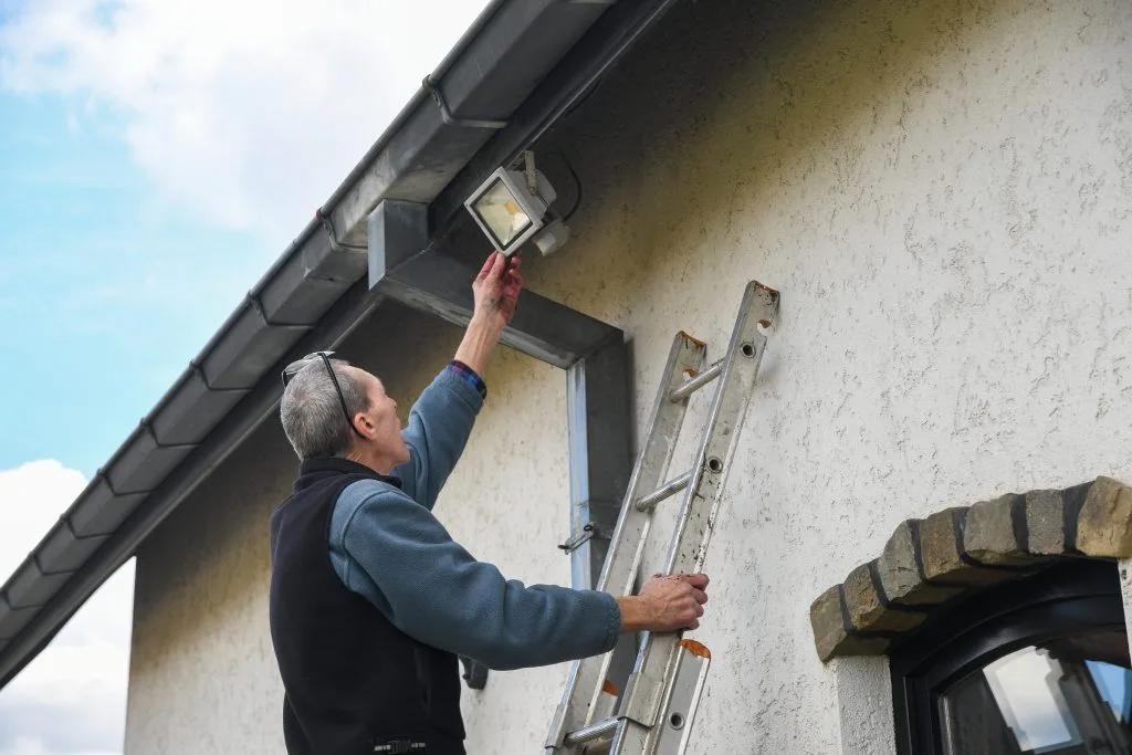 A man adjusts a light fixture on a wall while standing on a ladder